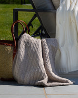 A chunky cable-knit throw blanket in a heather taupe -beige color draped over a black outdoor lounge chair next to a white knit blanket and a woven straw tote bag on a patio.
