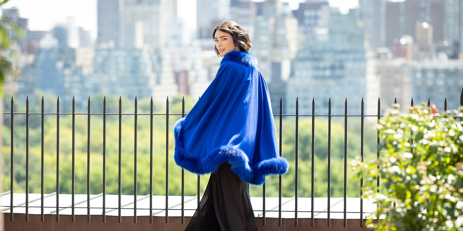 Woman wearing a blue fur-trimmed cashmere cape with a cityscape in the background