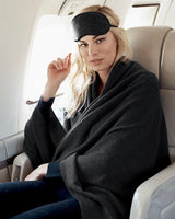 Woman sitting in an airplane with a sleep mask and blanket, Jersey Knit cashmere Travel Set; pouch, eye mask, socks, blanket in Charcoal