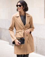Woman wearing a camel cashmere wool blend coat with sunglasses and a clutch against a light-colored wall.