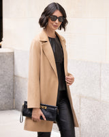 Woman wearing Bella Cashmere belted Coat in camel leather pants against a light-colored wall.
