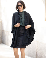 Woman wearing a Knitted Cashmere Cape in Black with ruffles, over an outfit with sunglasses, standing against a light wall.