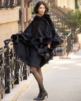 Woman wearing a Cervinia fur trimmed cashmere cape in black and fur trimmed leather gloves on a city street.