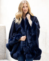 Woman wearing a fur-trimmed cashmere cape in navy against a light background