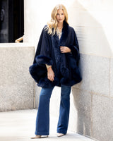 Woman wearing a cervinia fur-trimmed coat cape in navy against a light stone wall.
