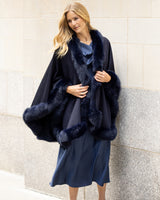 Woman wearing a formal dress with a fur-trimmed cape in navy against a light stone wall.