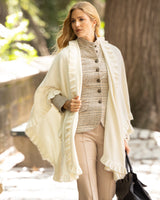 Woman wearing Knitted Cashmere Cape in Ivory with ruffles over a plaid shirt and plain pants, standing outdoors.