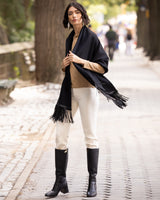 Woman wearing a Cashmere Wrap with Leather Fringe in black, beige sweater, white pants, and black boots on a street.