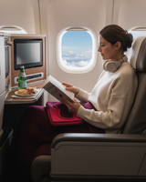 Woman reading a book on an airplane with burgundy cashmere blanket and sleep mask on lap with a view of clouds outside the window.