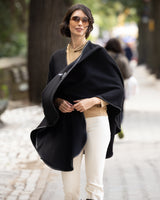 Woman wearing a reversible Cashmere Cape with Leather trim in Black/Black over a neutral sweater, and bright pants on a city street.