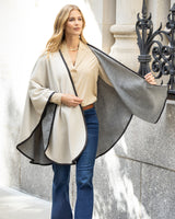Woman wearing a Reversible Cashmere Cape with Leather trim in Oat/Grey over a silk blouse and jeans, standing against a stone wall.