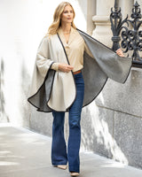 Woman wearing a Reversible Cashmere Cape with Leather trim in Oat/Grey over a blouse and jeans, standing in front of a stone wall.