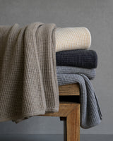 Stack of textured knit cashmere blankets in various colors on a wooden bench against a plain background