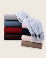 A stack of folded woolen scarves or throws in multiple colors, with a powder blue fringed throw draped over the top of the stack on a light beige background.