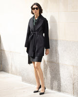 Woman wearing a Victoria cashmere belted coat in black and dress against a light stone wall.