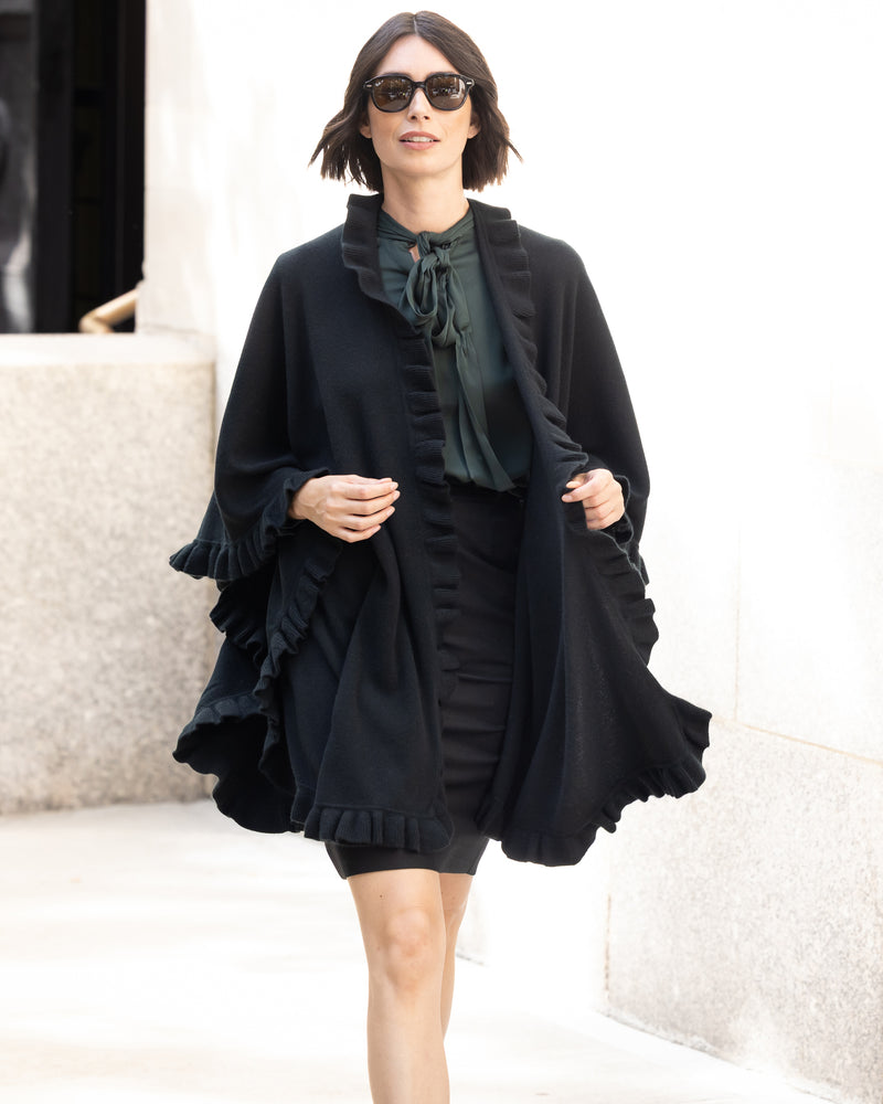 Woman wearing a Knitted Cashmere Cape in Black with ruffles, pencil skirt and blouse standing against a white background.