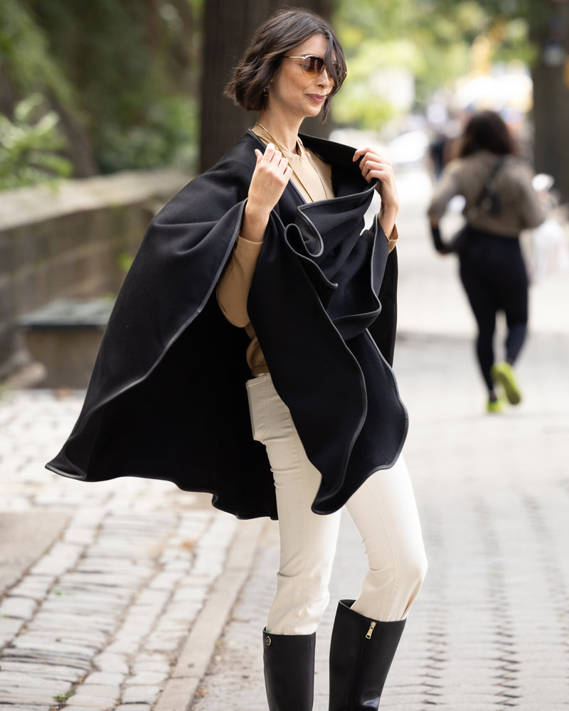 Woman wearing a reversible Cashmere Cape with Leather trim in Black/Black,  white pants, and black boots on a street.
