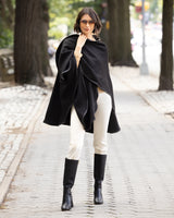 Woman wearing a reversible Cashmere Cape with Leather trim in Black/Black,  white pants, and black boots on a street.