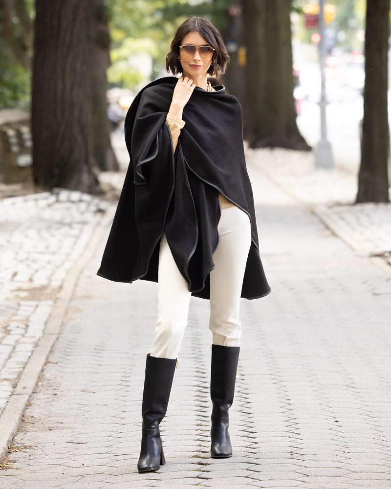 Woman wearing a reversible Cashmere Cape with Leather trim in Black/Black,  white pants, and black boots on a street.