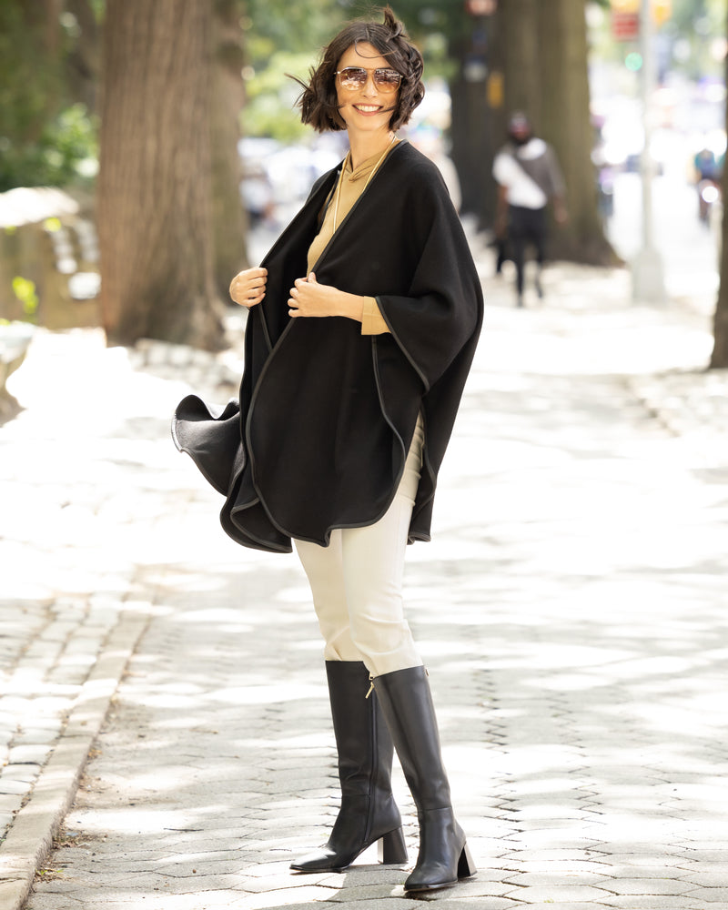 Woman wearing a reversible Cashmere Cape with Leather trim in Black/Black,  white skirt, and black knee-high boots on a city street.