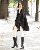 Woman wearing a reversible Cashmere Cape with Leather trim in Black/Black,  white pants, and black knee-high boots on a city street.