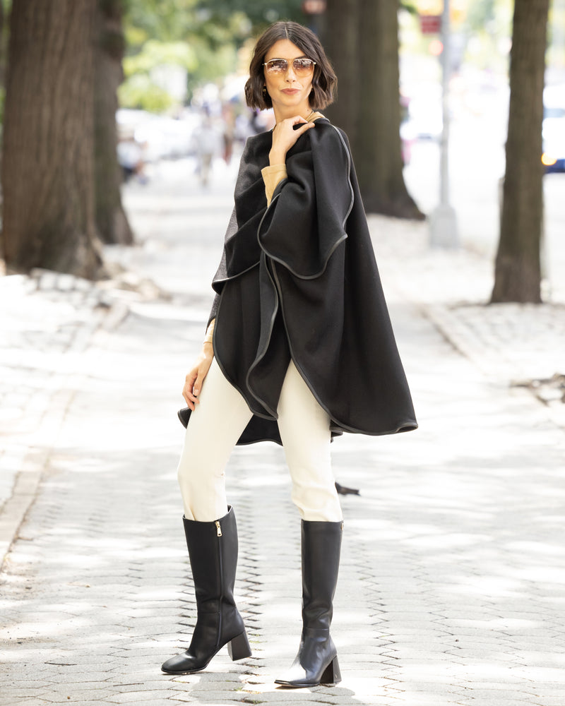 Woman wearing a reversible Cashmere Cape with Leather trim in Black/Black,  white pants, and black knee-high boots on a city street.
