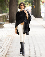 Woman wearing a reversible Cashmere Cape with Leather trim in Black/Black,  white pants, and black boots on a city street.