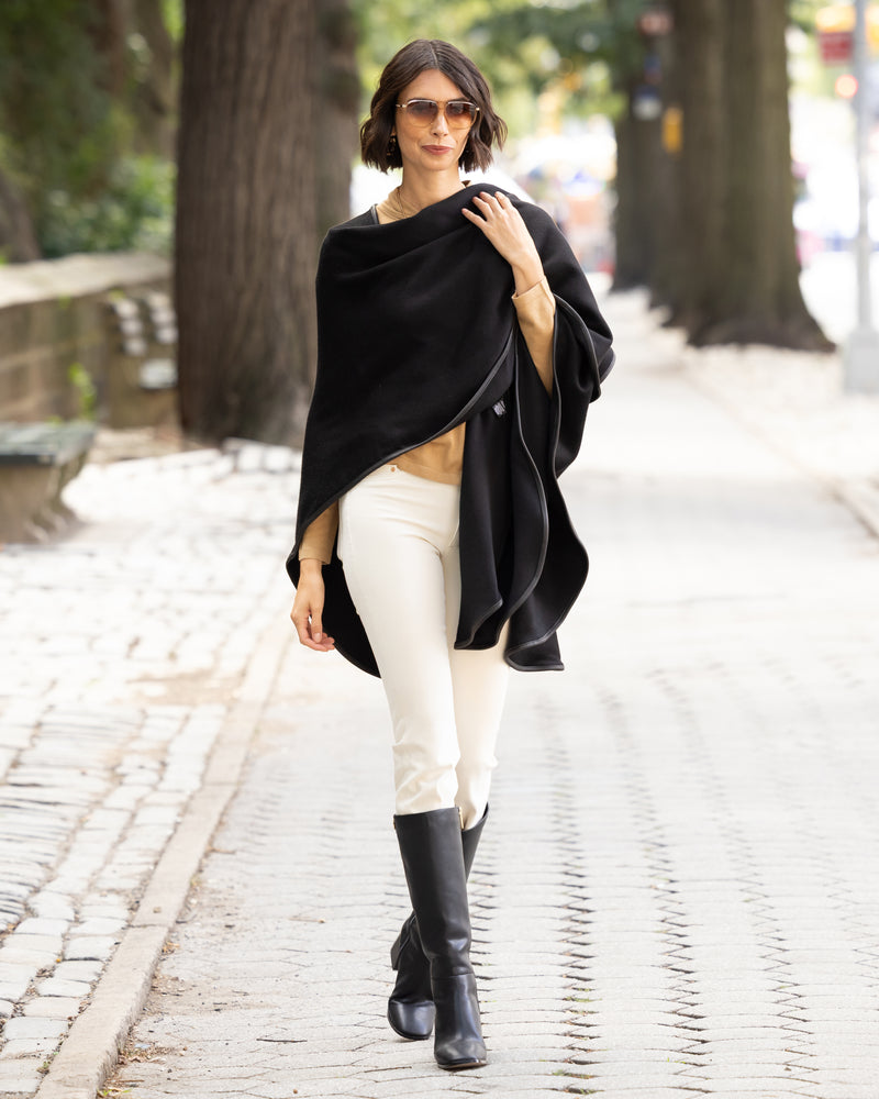 Woman wearing a reversible Cashmere Cape with Leather trim in Black/Black,  white pants, and black boots on a city street.