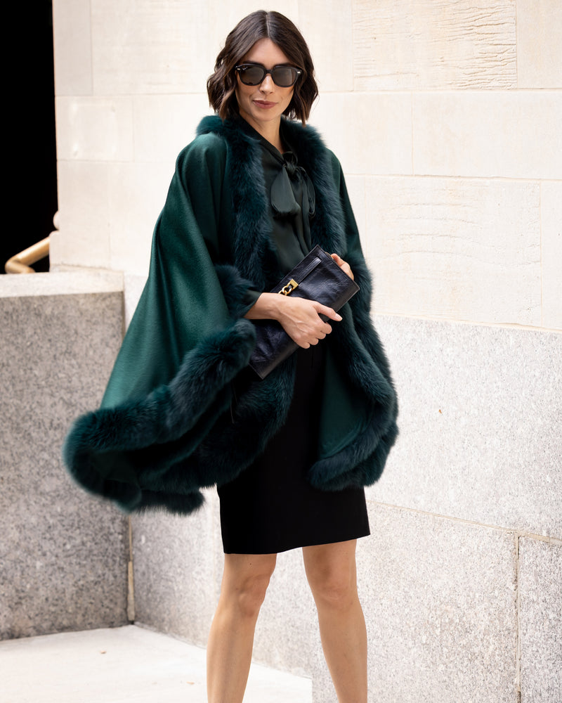 Woman wearing Fur Trimmed Cape in Emerald against a light stone wall.