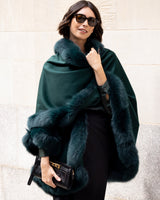 Woman wearing Fur Trimmed Cape in Emerald against a light wall.