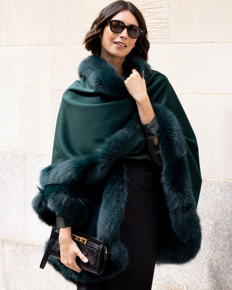 Woman wearing Fur Trimmed Cape in Emerald against a light wall.