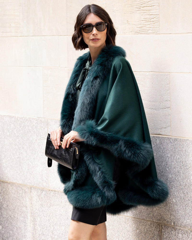 Woman wearing a Fur Trimmed Cape in Emerald against a stone wall.