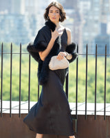 Woman in a strappy dress with Fur Trimmed Cashmere Shawl in black  holding a sequin clutch against a blurred cityscape background.