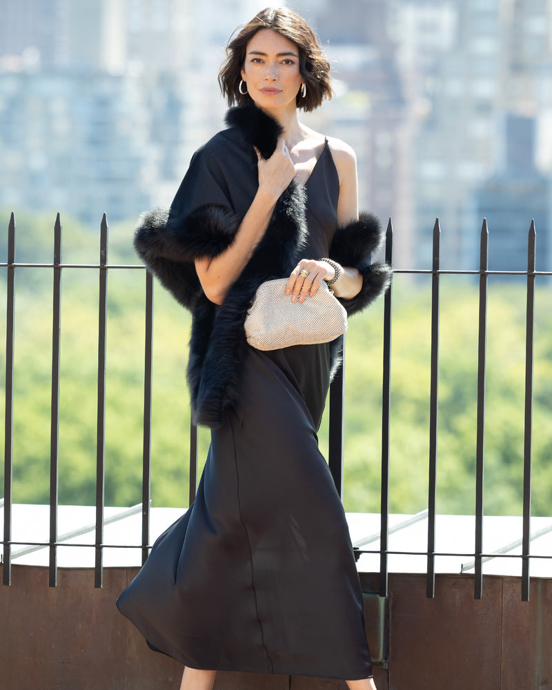 Woman in a strappy dress with Fur Trimmed Cashmere Shawl in black  holding a sequin clutch against a blurred cityscape background.