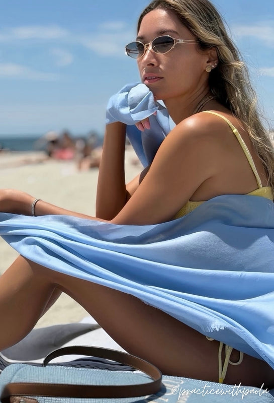 Woman sitting on a beach with a blue lightweight wrap and sunglasses, wearing a yellow bikini. @practicewithpaola