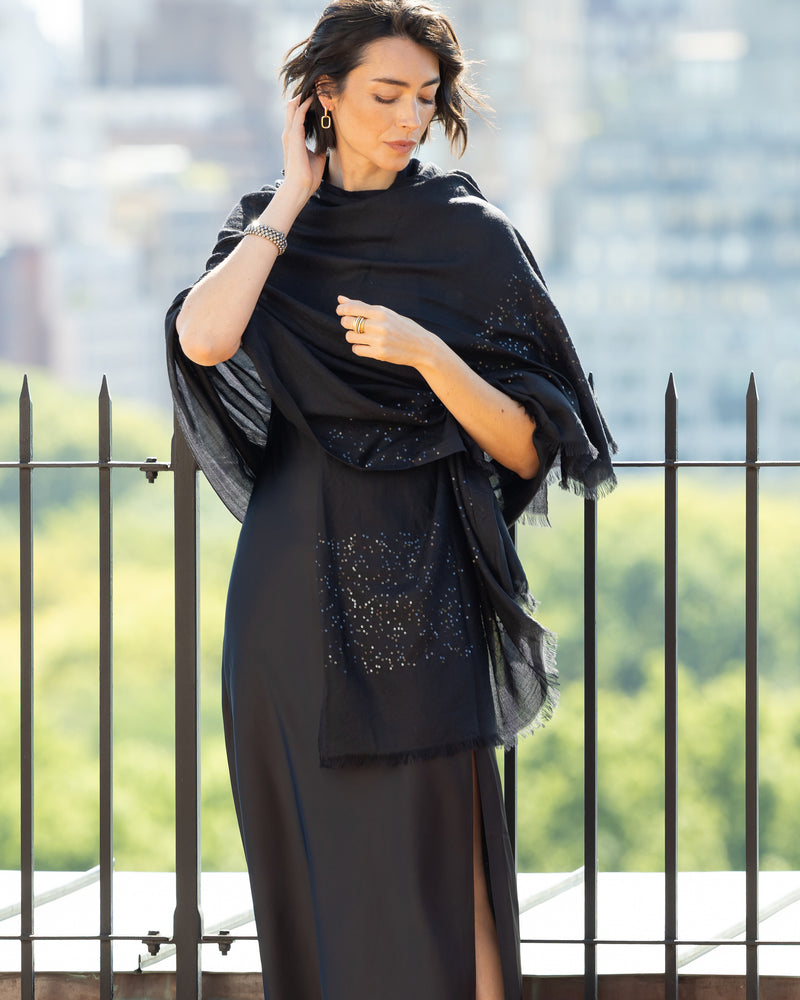 Woman wearing a dress with a Aura Lightweight Cashmere Sequin Wrap in black against a blurred outdoor background