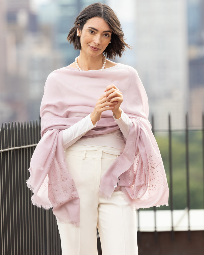 Woman wearing Aura Lightweight Cashmere Sequin Wrap in light pink with a blurred cityscape background