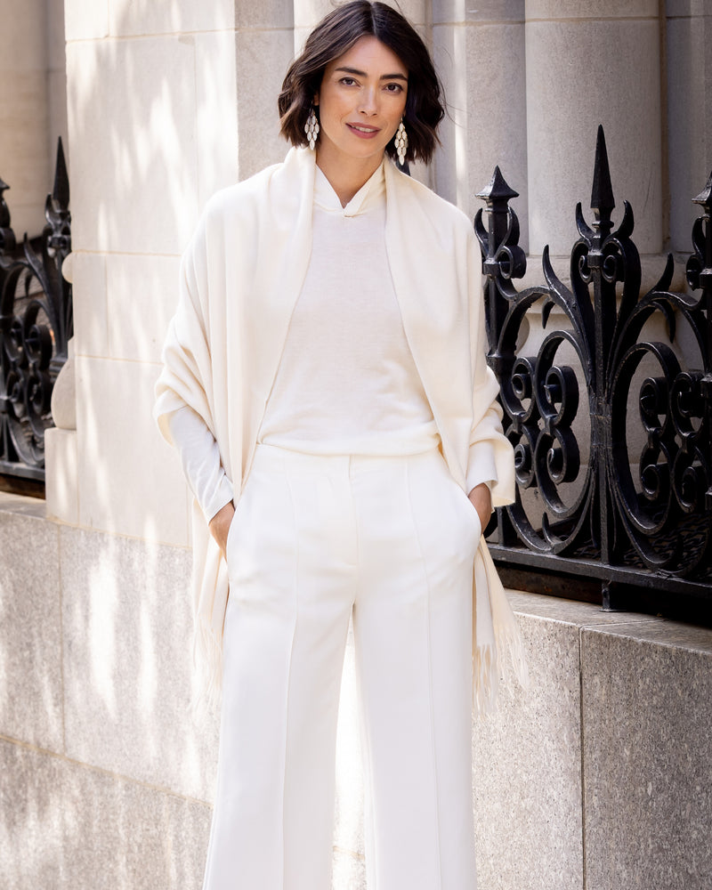 Woman wearing Cashmere water weave wrap with fringe in ivory standing against a stone wall with decorative metalwork.