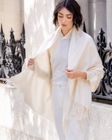 Woman wearing Cashmere water weave wrap with fringe in ivory over a light colored outfit, standing in front of decorative metal railings.