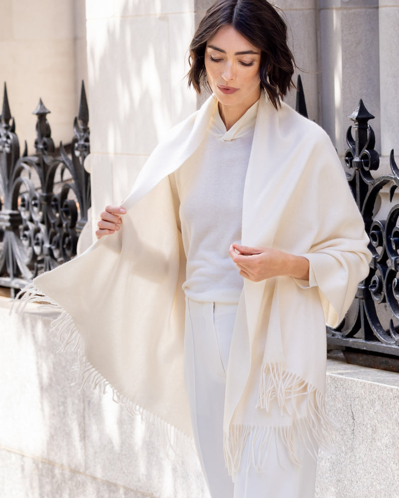 Woman wearing Cashmere water weave wrap with fringe in ivory over a light colored outfit, standing in front of decorative metal railings.