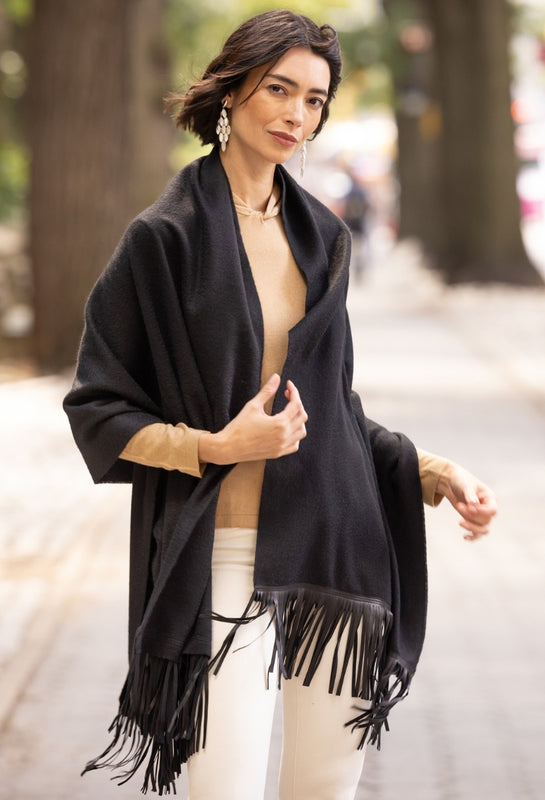 A woman wearing a black fringed wrap over a beige top and white pants, standing on a blurred outdoor street with trees in the background.