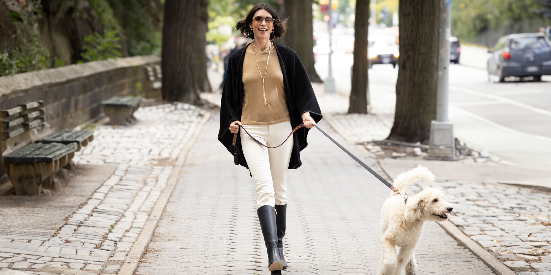 Woman walking in a city park side wearing a cashmere cape with leather trim while walking a dog