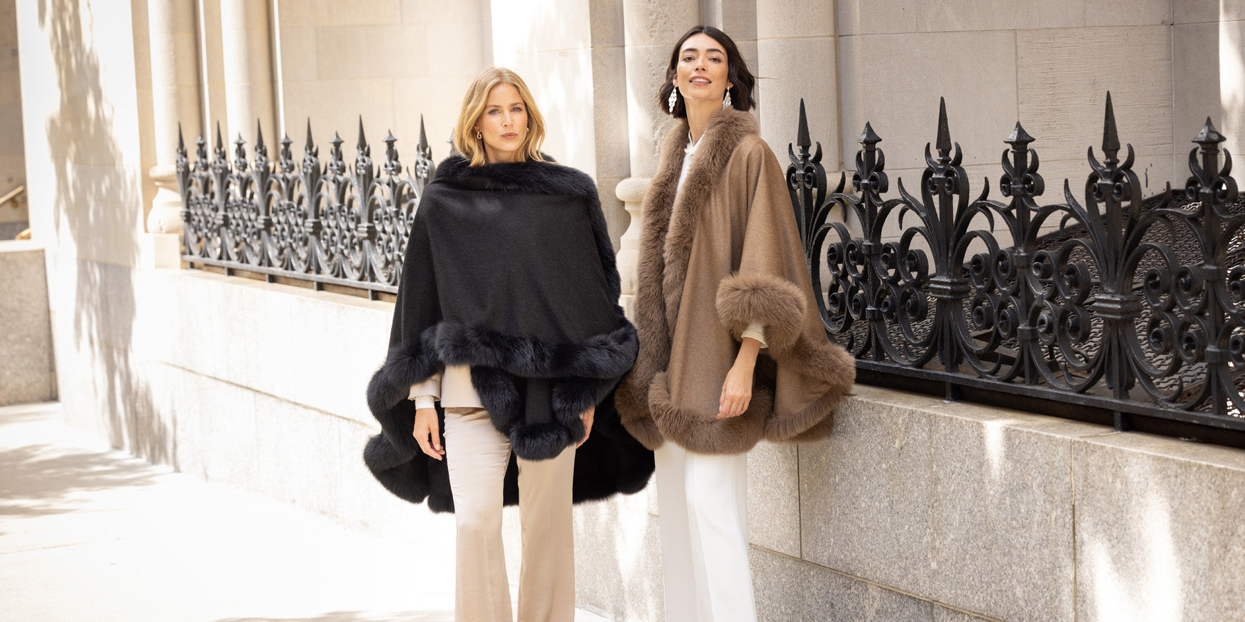 Two women wearing fur-trimmed capes standing in front of a decorative metal railing.