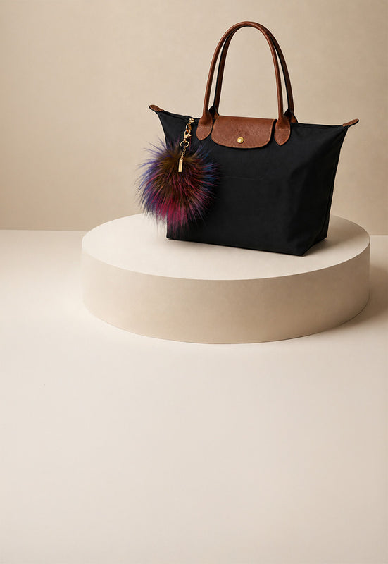 A black canvas tote bag with brown leather handles and trim, displayed on a round beige pedestal. It features a colorful purple and pink faux-fur pom-pom charm with gold hardware.