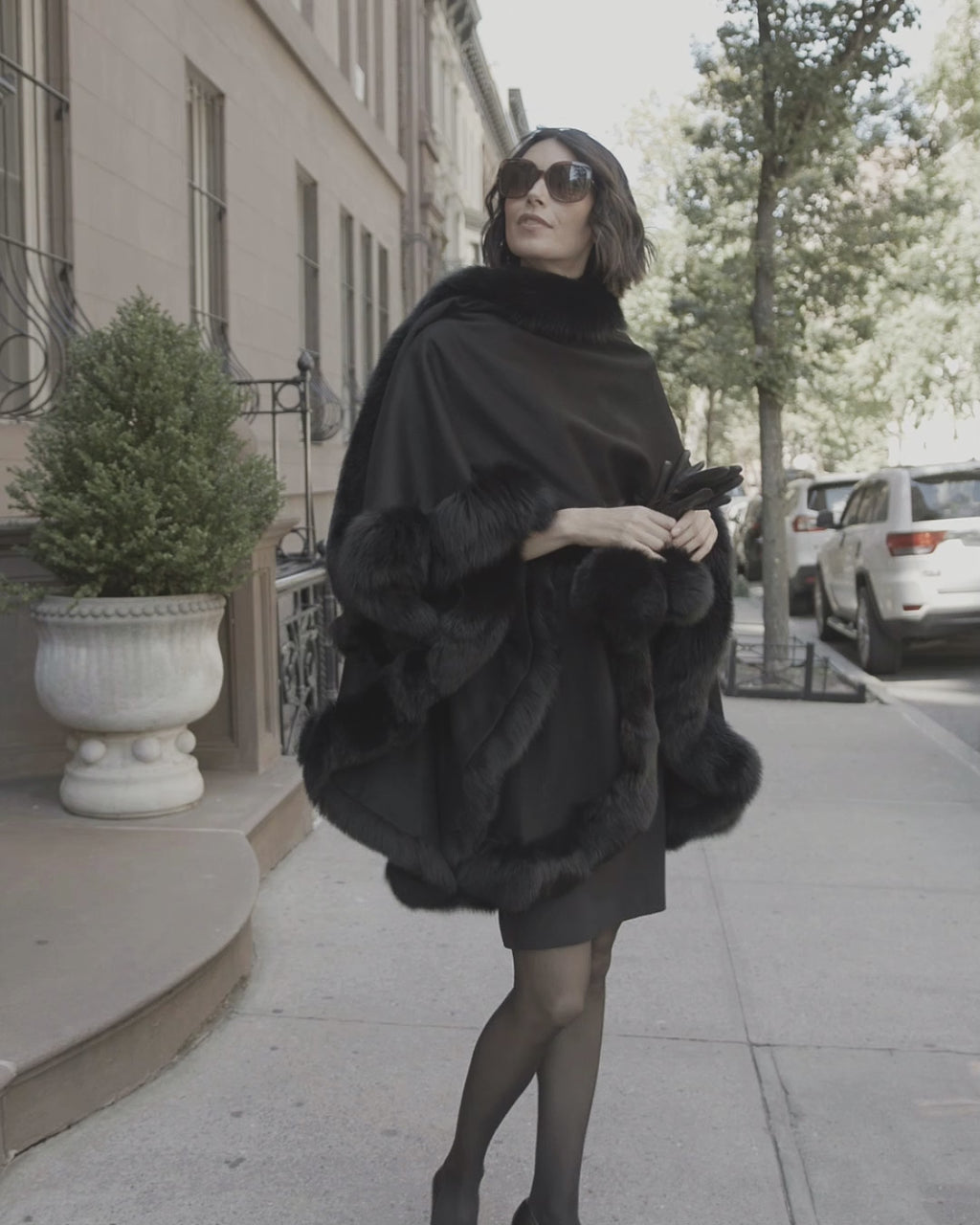 Woman waling wearing sunglasses and sofia cashmere fur trimmed cashmere cape in black