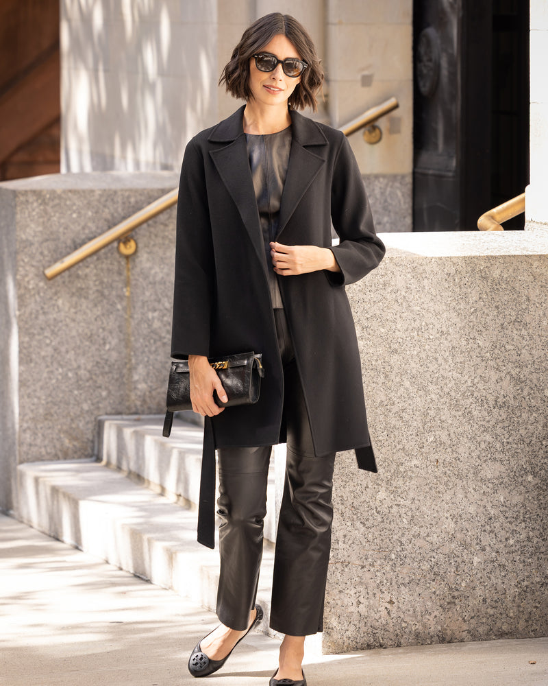 Woman in Bella Cashmere belted Coat in black and pants standing outdoors near a building.