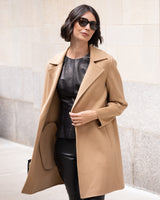 Woman wearing Bella Cashmere belted Coat in camel and sunglasses against a light-colored wall.