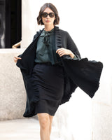 Woman wearing Knitted Cashmere Cape in Black with ruffles, with a blouse, skirt and sunglasses, standing against a neutral background.