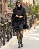 Woman wearing a Cervinia fur trimmed cashmere cape in black and sunglasses on a city street.