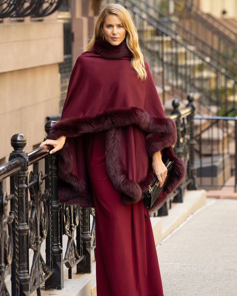 Woman wearing a fur trimmed cashmere cape in burgundy standing on a city street.